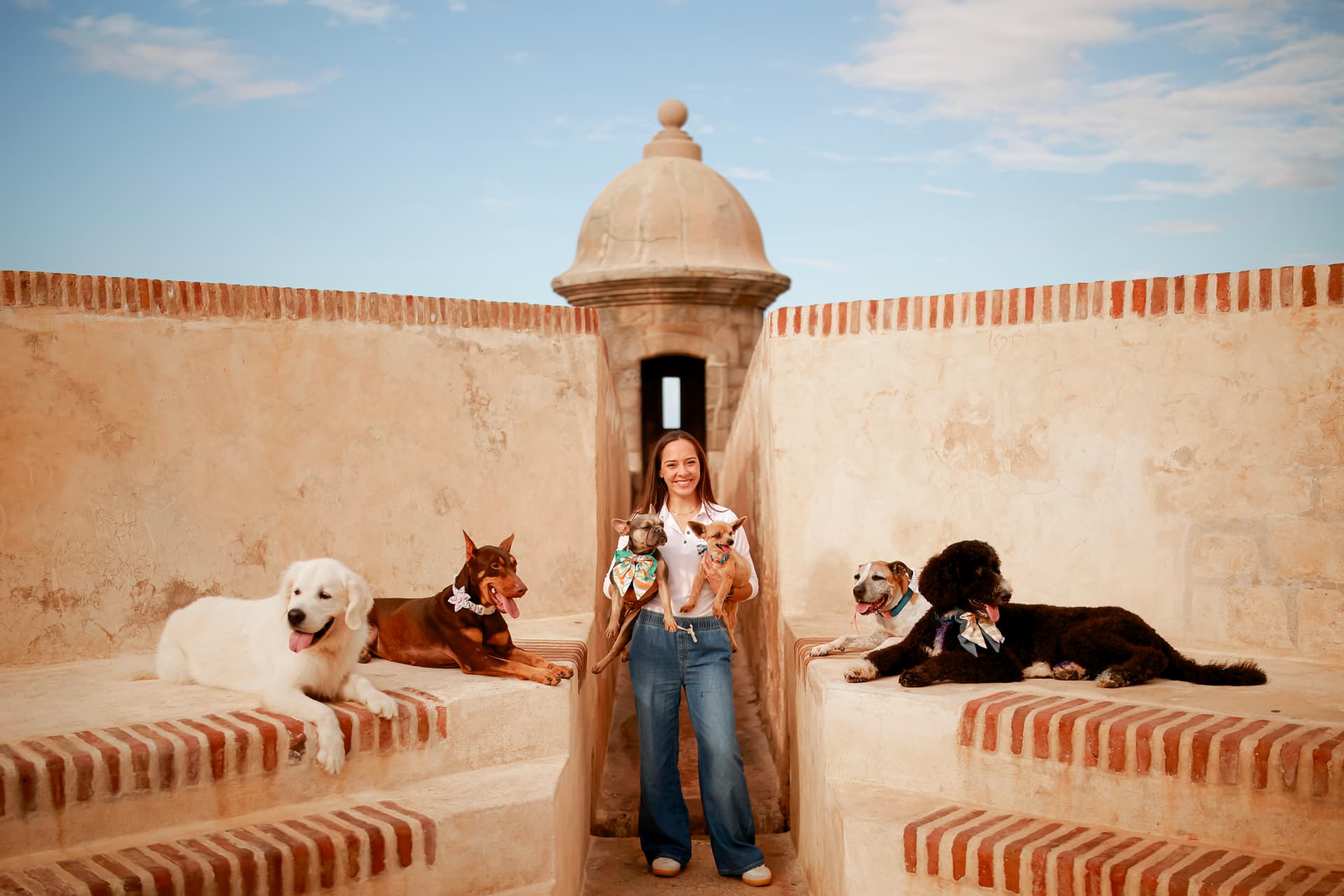 Trainer Anayla Fontán Núñez smiling at a historic Puerto Rico fort, surrounded by six trained dogs of different breeds resting calmly on the stone steps around her